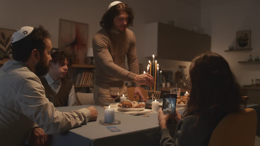 Medium shot of widowed middle-aged Jewish man in kippah celebrating Hanukkah at home with his children, eldest son lighting up candles on menorah and young daughter filming on smartphone Royalty-Free Stock Footage #1111584493