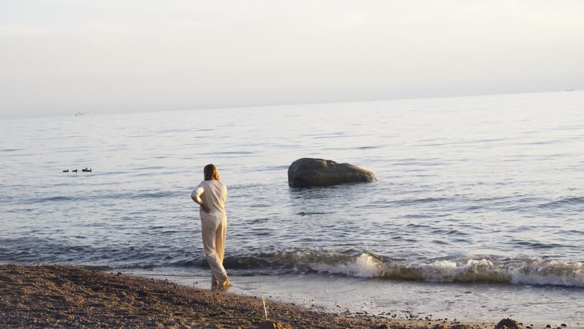 A woman of European appearance, 40 years old, in a white suit, walks along the seashore in the water barefoot, enjoying the sunset. Holidays at sea.