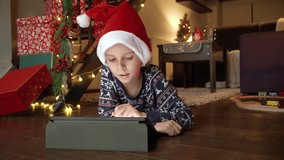 Happy smiling boy wearing Santa's hat lying on floor in living room and playing on tablet computer. Winter holidays, celebrations and party