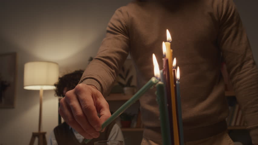 Close-up shot of hand of unrecognizable young man holding shammash and lighting up all candles on hanukkiah on final day of Hanukkah holiday, and young boy in kippah sitting in background Royalty-Free Stock Footage #1111587079