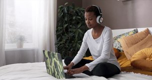 Curious, happy African American lady having video call conference interview with boss while sitting on bed wearing new headphone discussing new projects ideas.