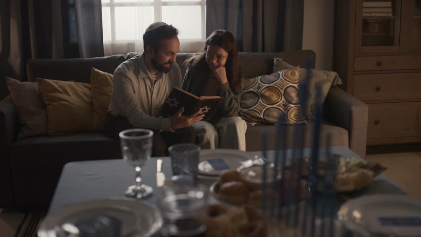 Full shot of bearded Jewish man in kippah sitting on couch with young girl, holding Torah book, reading and discussing holy scripture, and festive table with hanukkiah in blurred foreground Royalty-Free Stock Footage #1111588709