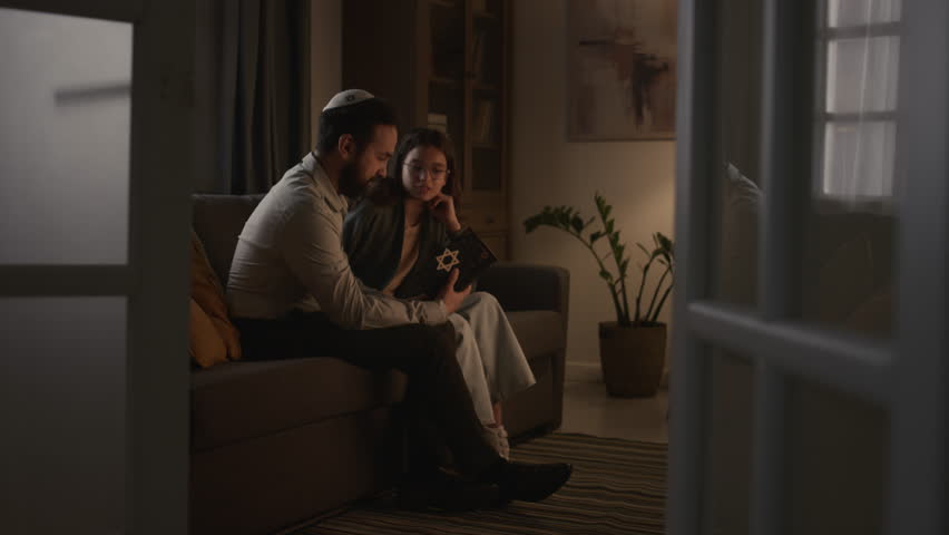 Full shot of middle-aged Jewish father in kippah and teenage daughter sitting together on couch at home, man holding holy Torah book, talking, asking questions, pointing and reading religious text Royalty-Free Stock Footage #1111588857