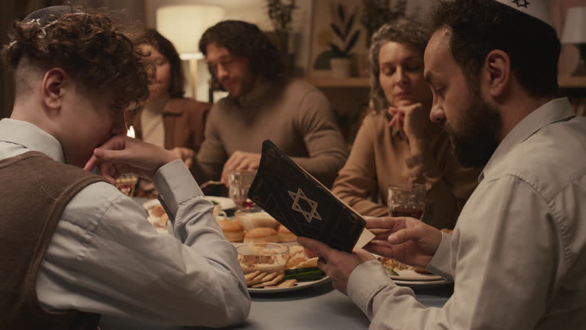 Medium shot of Jewish man in kippah sitting at Hanukkah dinner with family and friends, reciting solemn passages from holy Torah book with star of David on cover, and people sitting and listening Royalty-Free Stock Footage #1111589307