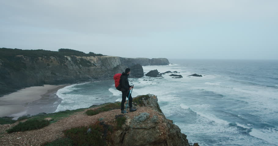 Wide open shot of young hispanic man look into distance over ocean and cliffs, stand on edge of rock on end point of pilgrim trail or path. Hiking and exploring wild coastline | Shutterstock HD Video #1111589503