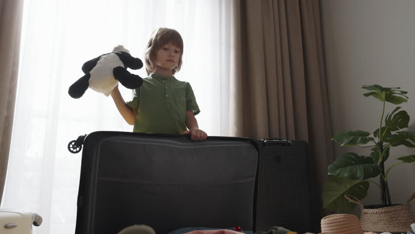 Boy throws another toy into his overfilled and disorderly suitcase. Then he climbs onto it with his feet and jumps on it, trying to close. Vacation dreams chaotic travel preparations, app advertising | Shutterstock HD Video #1111591697