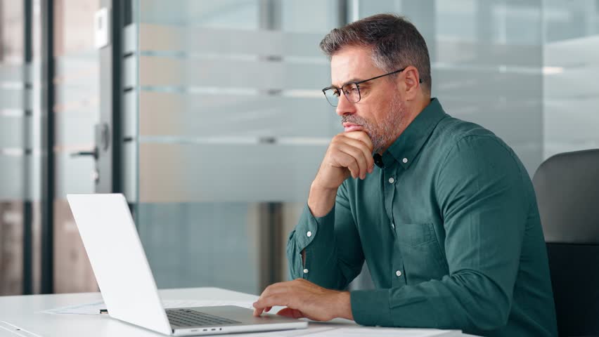 Busy middle aged professional business man executive investor using computer working at desk. Male manager expert looking at laptop thinking on online finance market analysis, elearning in office.
