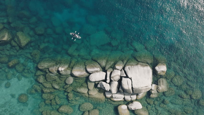 Sun light refracting of transparent water around rocky island. Couple kayaking together in pristine blue lagoon in lake Tahoe 4K. Drone view tourists on weekend adventure paddling in clear kayak 4K
