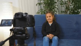 Happy blogger girl talking to camera while sitting on sofa in room. Child blogger sits at home and talks to audience live. Child video blogger. Girl behind scenes is recording video blog on camera.