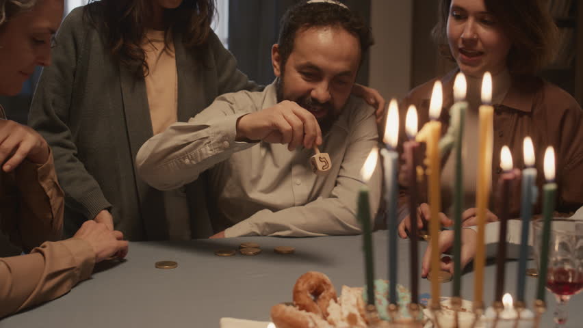Medium shot of middle-aged Jewish man with beard, in kippah, playing fun traditional game with family at Hanukkah dinner, spinning dreidel, cheering, laughing and betting gelt coins Royalty-Free Stock Footage #1111596833