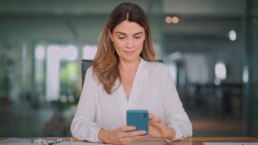 Happy mid aged 40s business woman holding phone using cellphone in office. Smiling mature professional businesswoman executive using smartphone cell mobile apps on cellphone working sitting at desk. | Shutterstock HD Video #1111597263