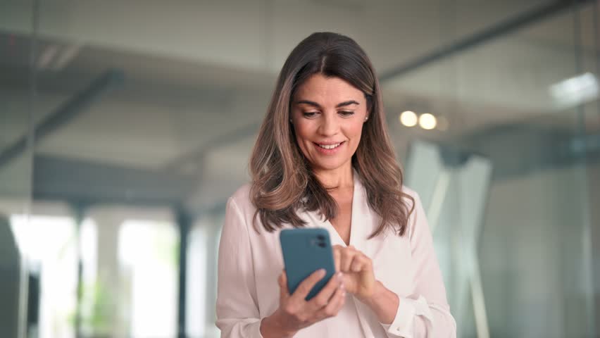 Mid aged mature happy professional business woman using mobile applications on phone at work standing at lobby, businesswoman entrepreneur holding smartphone reading messages on cellphone in office. | Shutterstock HD Video #1111597267
