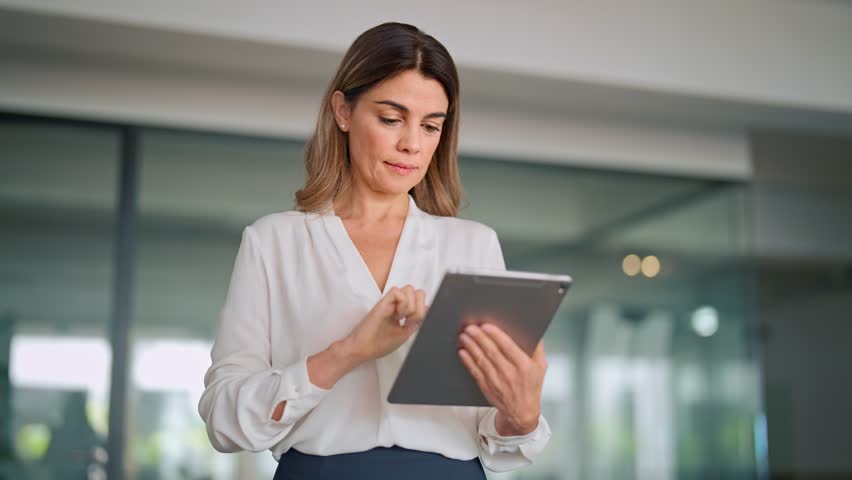Mature busy latin business woman financial manager using digital tablet working in office. Middle aged professional businesswoman executive holding tab technology device standing at work. | Shutterstock HD Video #1111597271