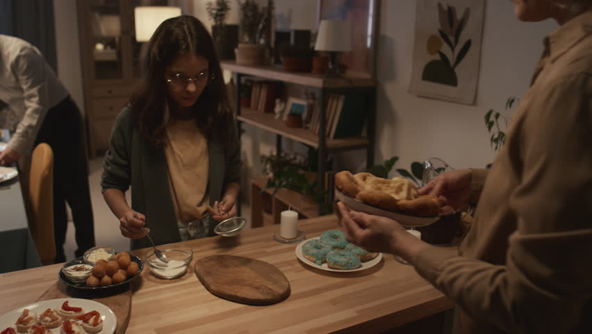 Medium shot of Jewish people preparing for Hanukkah dinner at home - daughter dusting sufganiyot with sugar powder, mom watching, father in kippah taking dish and carrying away to put on table Royalty-Free Stock Footage #1111601493