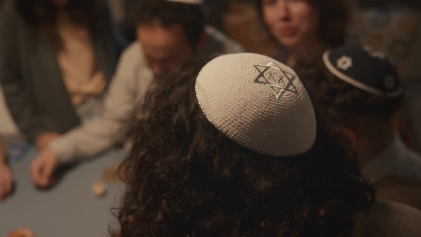 Close-up selective focus shot of Jewish traditional headwear, kippah decorated with star of David, on head of young anonymous male playing dreidel game with family and friends at Hanukkah party Royalty-Free Stock Footage #1111602101