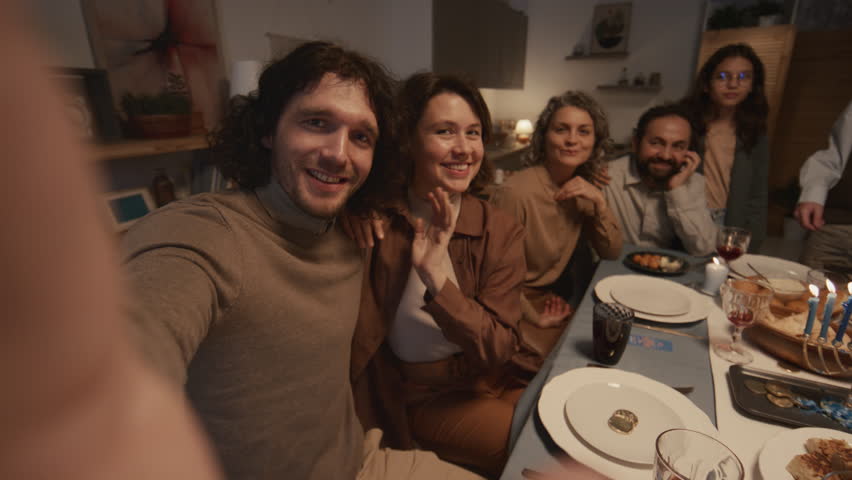 Medium selfie shot of young Jewish man in kippah sitting at dinner table with wife, parents and teenage siblings, holding smartphone and preparing to take photo of happy family gathering Royalty-Free Stock Footage #1111602817