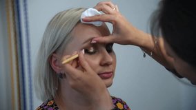 Close-up view of make-up artist applying golden glittering powder eyeshadow makeup using brush on adult blond woman's eyelids in beauty salon. Soft focus. Real time handheld video. Make-up theme.