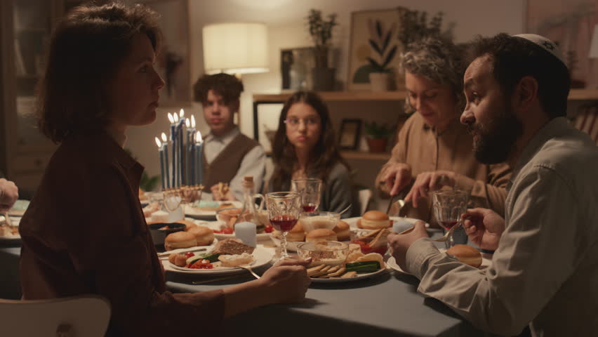 Medium shot of Jewish family sitting around festive table and enjoying traditional foods at Hanukkah dinner, dad in kippah talking to female relative or friend, then both laughing Royalty-Free Stock Footage #1111609343