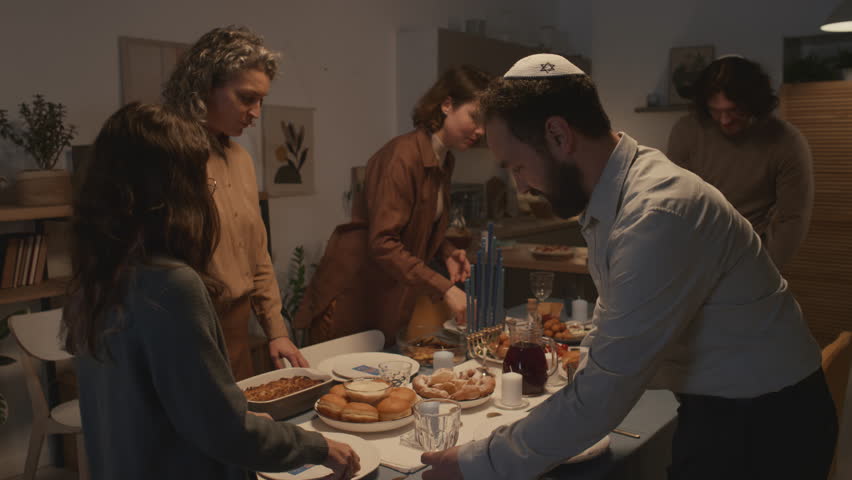 Medium shot of Jewish family of five, men wearing kippah, getting ready for Hanukkah dinner together at home, putting cutlery and food on table, chatting and smiling Royalty-Free Stock Footage #1111612115
