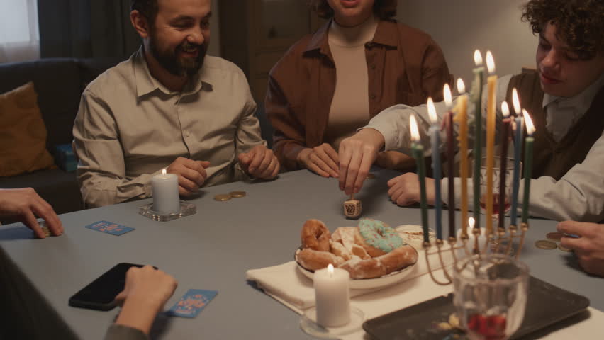 Medium shot of teenage Jewish boy wit curly hair, in kippah playing dreidel with family during Hanukkah feast, spinning, getting Hebrew letter Hei, meaning half, taking gelt coins, and all laughing Royalty-Free Stock Footage #1111612421