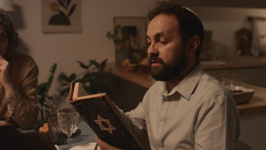Medium shot of middle-aged Jewish man in kippah sitting at festive dinner with family, celebrating Hanukkah in candlelight, reading passages from holy Torah with star of David on cover Royalty-Free Stock Footage #1111615431