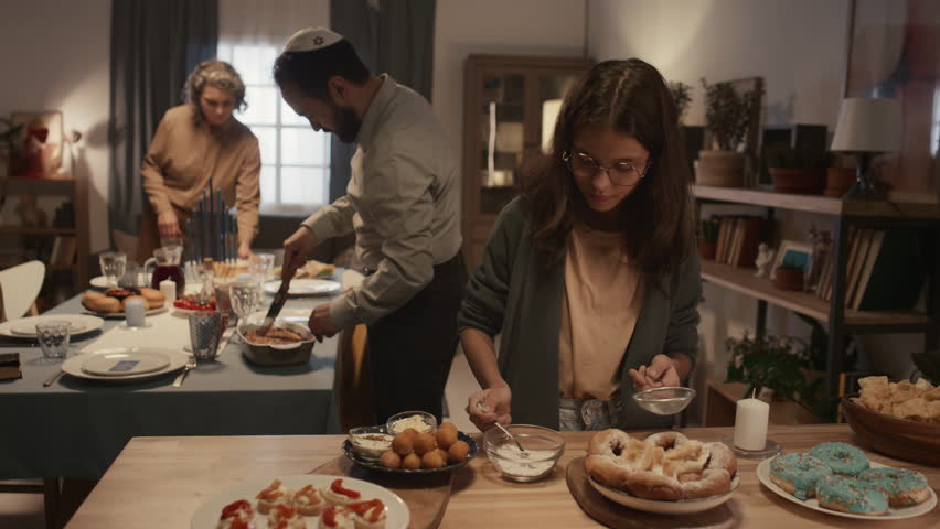 Medium shot of teenage Jewish girl sprinkling icing sugar onto sufganiyot and fritters, while mother and father in kippah are cutting kugel and laying out cutlery on Hanukkah table in background Royalty-Free Stock Footage #1111615839