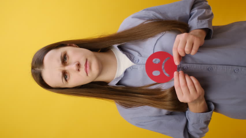 Vertical video of serious young caucasian woman holding red unsatisfied sad face emoticon, posing isolated over plain yellow color background wall in studio. Concept angry, stressed, negative emotion | Shutterstock HD Video #1111615967