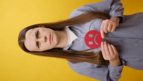 Vertical video of serious young caucasian woman holding red unsatisfied sad face emoticon, posing isolated over plain yellow color background wall in studio. Concept angry, stressed, negative emotion - Powered by Shutterstock - Get 15% off with code: PIKWIZARD15