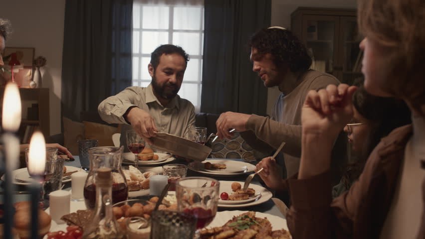 Medium shot of cheerful Jewish family eating dinner together on final day of Hanukkah, smiling, chatting, father in kippah holding tray, son taking out piece of kugel and laughing Royalty-Free Stock Footage #1111617383