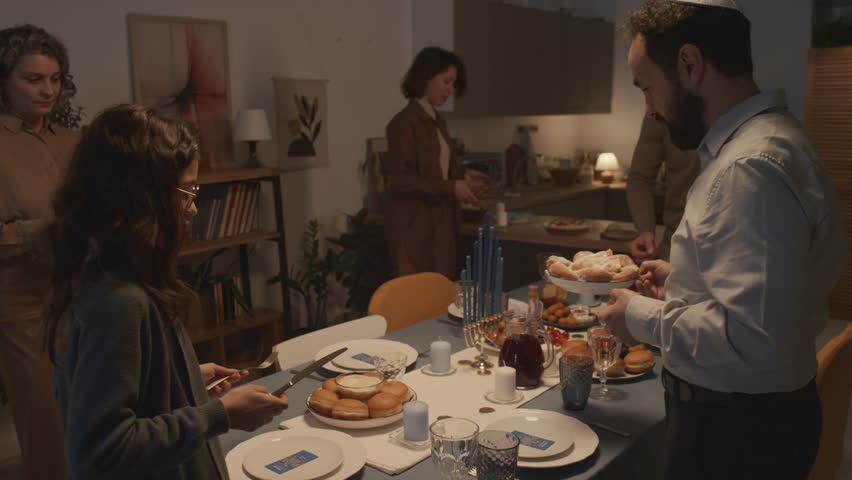 Medium shot of Jewish family of five, men wearing kippah, getting ready for Hanukkah dinner together at home, putting cutlery and food on table, chatting and smiling Royalty-Free Stock Footage #1111617553