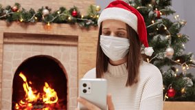 Adult woman with brown hair wearing Santa Claus hat and medical mask sitting in decorated living room with Christmas tree and burning fireplace talking via smartphone social distance virus disease. - Powered by Shutterstock - Get 15% off with code: PIKWIZARD15