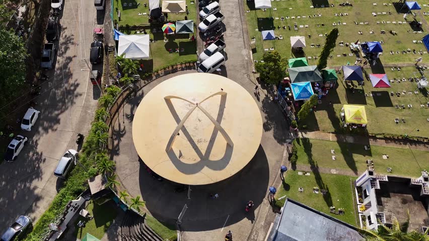 Aerial drone footage of Memorial Park in Surigao City - Philippines, on All Souls Day, showing cars parked and gazebos on a field surrounded by lush greenery. Daytime.