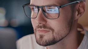 Young guy looking at screen with glasses reflection. Web designer browsing stock photos content he could buy. Concentrated man in eyeglasses scrolling social networks