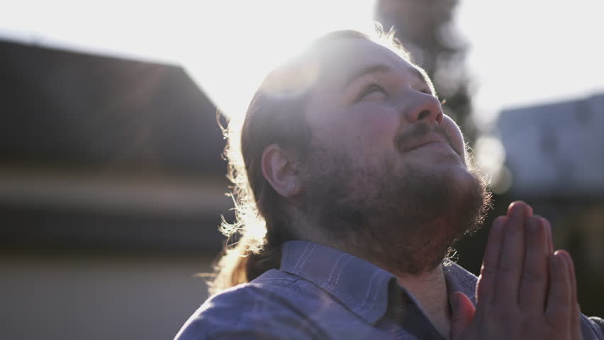 Happy man praying to God with hands clenched in prayer standing outdoors in sunlight having GRATITUDE