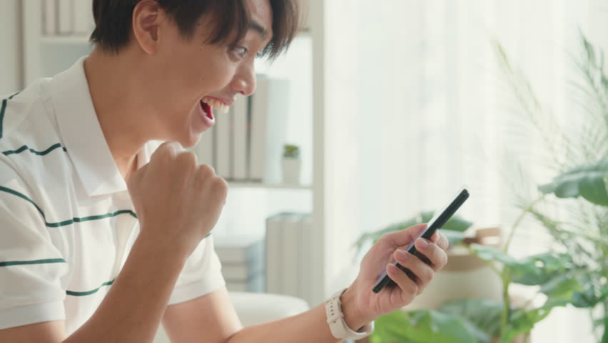 Close up youth Asia male with casual cloth sit on sofa looking at smartphone feel excited with football match score at living room in cozy house. Online shopping, Lifestyle leisure at home concept. | Shutterstock HD Video #1111634889