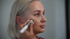 Close-up view of make-up artist applying cream makeup foundation using brush on adult blond woman's face in beauty salon. Soft focus. Real time handheld video. Make-up theme.