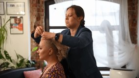 Side view of female hairdresser making new coiffure for adult blond haired woman using hair clip (or barrette) in hair salon. Soft focus. Real time handheld video. Beauty industry theme.