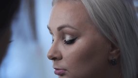Close-up view of make-up artist applying mascara using wand applicator on eyelashes of adult blond woman in beauty salon. Soft focus. Real time handheld video. Make-up and beauty theme.