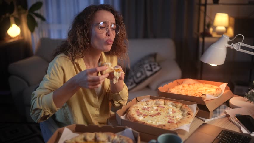 A young, curly-haired freelance woman dressed in a yellow shirt works at home at night. Her desk is lined with boxes of pizza and she enjoys the taste of it while working | Shutterstock HD Video #1111635071
