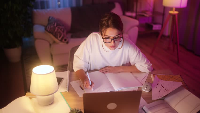 Concentrated pretty girl preparing for exam at university studying at home workplace Busy student make notes while looking at laptop computer screen indoors Distance remote education | Shutterstock HD Video #1111635079