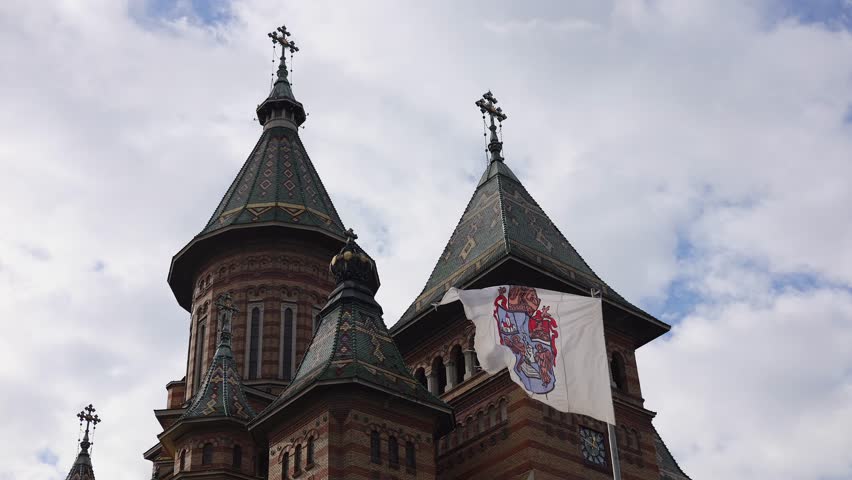 The flag of Timisoara city waving against cloudy sky and Orthodox Cathedral Three Holy Hierarchs. Travel to Romania.
