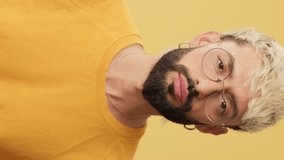 Vertical video, Close up, guy with glasses, dressed in yellow T-shirt, looking at camera with big eyes, says wow, covers his mouth with his hands, isolated on yellow background in studio