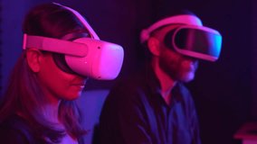 Bearded caucasian man and young asian woman sitting on the sofa at home and using Virtual Reality headset with joysticks playing VR video games 4K.