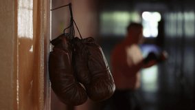 The shot shows a close up of a doorjamb with a rusty nail nailed to it on which boxing gloves hang. In the background a man in boxing gloves stands in a rack and hones his punches before a fight.