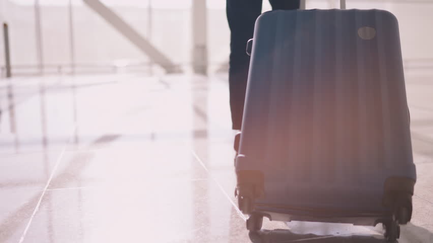 Close-up of blue suitcase, belonging to traveler glides along glossy airport floor. Concept of constant movement and pace of modern travel. Walking with suitcase in terminal to departure