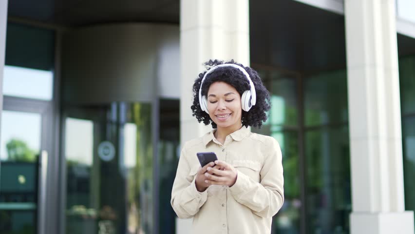 Happy joyful african american female student listens to music on headphones walking on campus space near university building. Smiling black woman turns on the playlist and enjoys song on smartphone