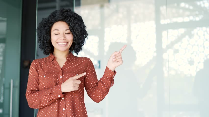 Happy young african american female employee pointing with finger at empty space for writing text standing in office. Black woman looks at camera. Template for advertising. Copy space for promotion
