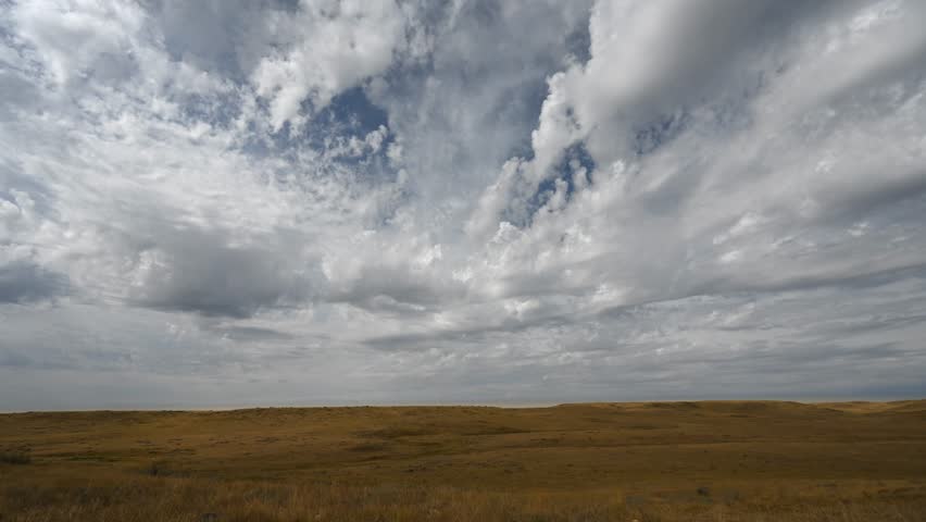 Time lapse of spectacular puffy white summer clouds moving towards the viewer over a dry prairie setting. Sunlight moves across the dry grass highlighting the small hills.
