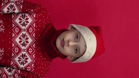 portrait of a surprised Caucasian boy 9 years old wearing a santa hat in a sweater and looking at the camera. vertical video. Christmas concept, gifts for children. surprise, children's emotions