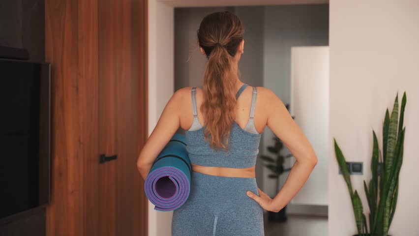Young happy woman going to do sports at home back rear view, holds karemat mat in hands, sportswear suit blue top leggings. modern room interior. aerobics sport fit yoga workout domestic fitness 4k | Shutterstock HD Video #1111662095